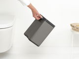 Grauer Abfallbehälter Bo 4L, seitlich gehalten von einer Hand, neben einer Toilette und einem Tisch mit Handtüchern.