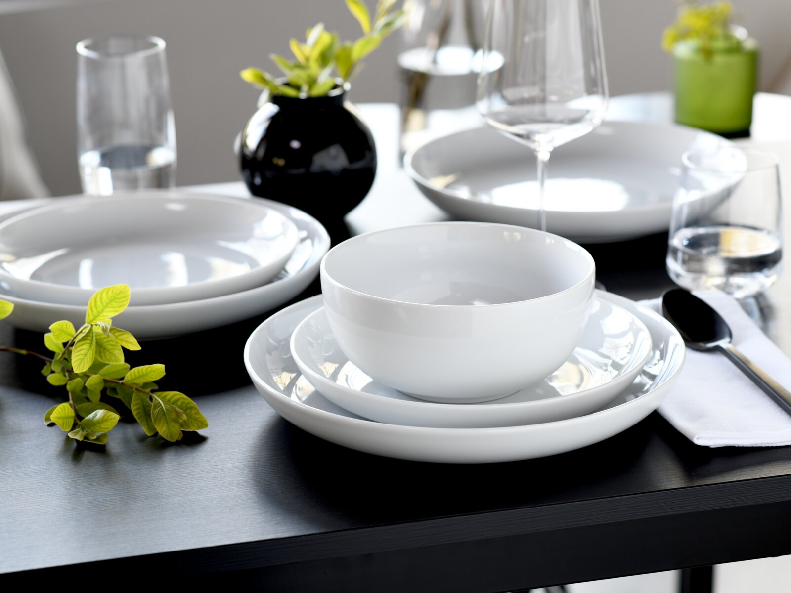 Modern gedeckter Esstisch mit weißem Porzellan-Geschirrset (Schüssel und Teller) auf dunklem Tisch, dazu Weinglas, Wassergläser, Besteck und kleine Pflanzendeko, aufgenommen aus leicht schräger Vorderansicht.