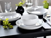 Modern gedeckter Esstisch mit weißem Porzellan-Geschirrset (Schüssel und Teller) auf dunklem Tisch, dazu Weinglas, Wassergläser, Besteck und kleine Pflanzendeko, aufgenommen aus leicht schräger Vorderansicht.