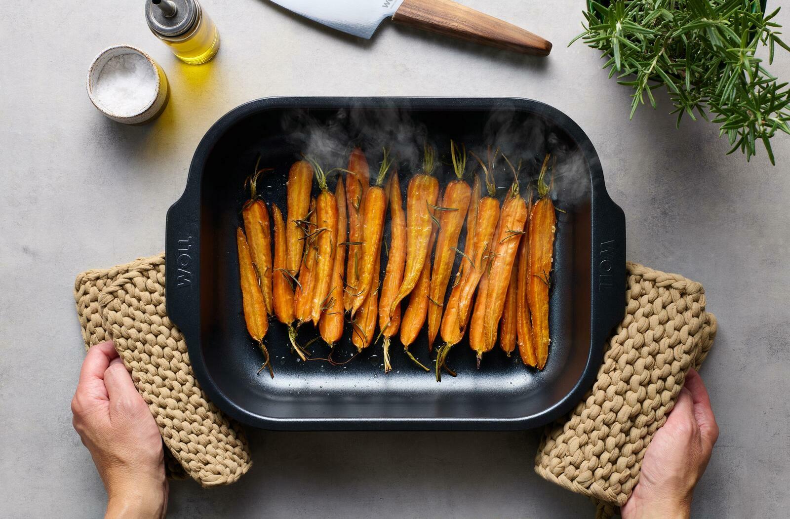 Obenansicht einer Bratreine mit gebackenen Karotten, gehalten von Händen mit Topflappen, umgeben von Salz, Öl und einem Messer.