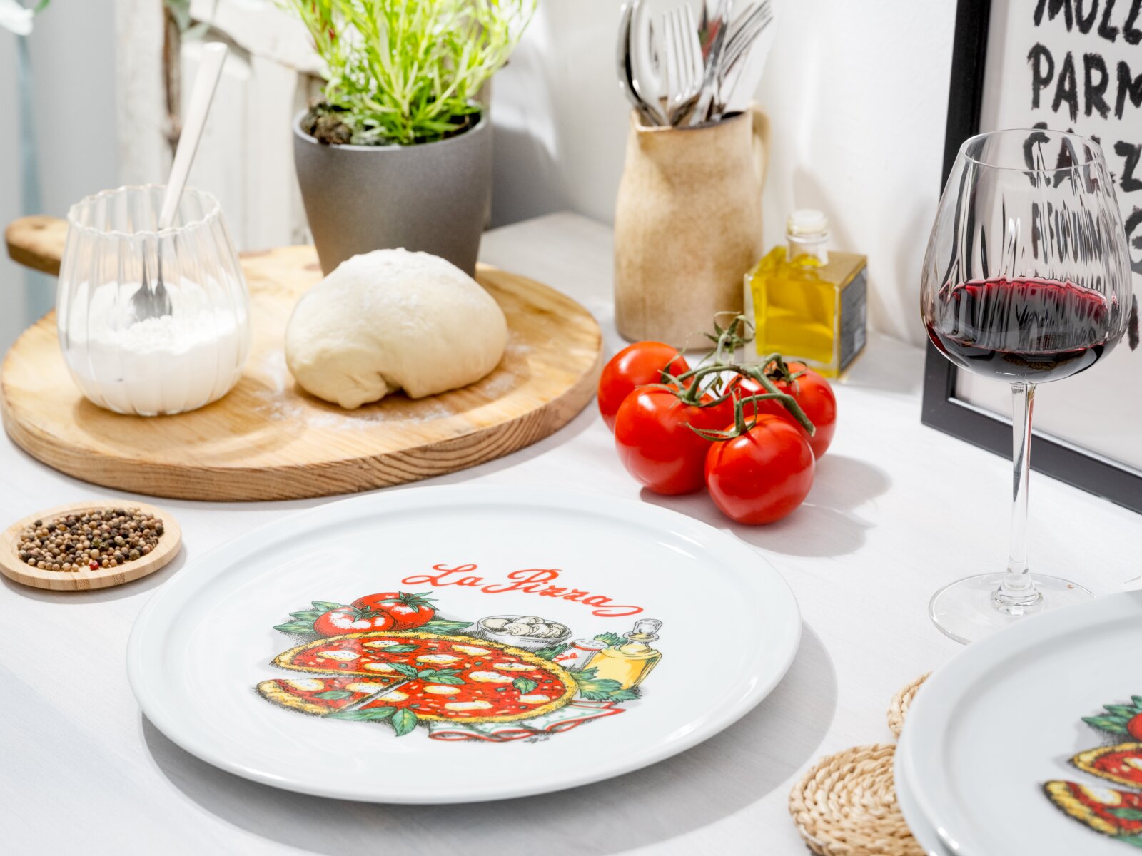 Weißer Teller mit Pizza-Motiv und Schriftzug „La Pizza“ auf gedecktem Küchentisch, umgeben von Teig, Tomaten, Olivenöl und Rotweinglas, fotografiert aus leicht schräger Seitenansicht von oben.