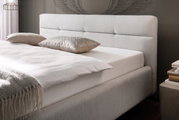 Zijaanzicht van een elegant gestoffeerd bed met lichte bekleding, decoratieve kussens en een gezellige deken.