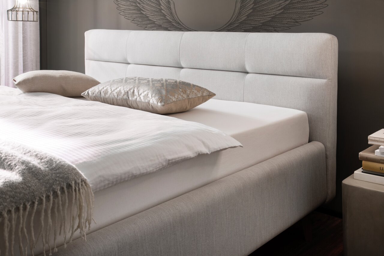 Zijaanzicht van een elegant gestoffeerd bed met lichte bekleding, decoratieve kussens en een gezellige deken.