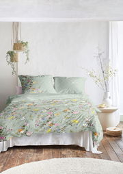 Bettwäsche-Set in sanftem Grün mit buntem Blumenmuster, auf einem Bett in einem hellen Raum mit Holzfußboden und Pflanzen, aus der Frontalperspektive.