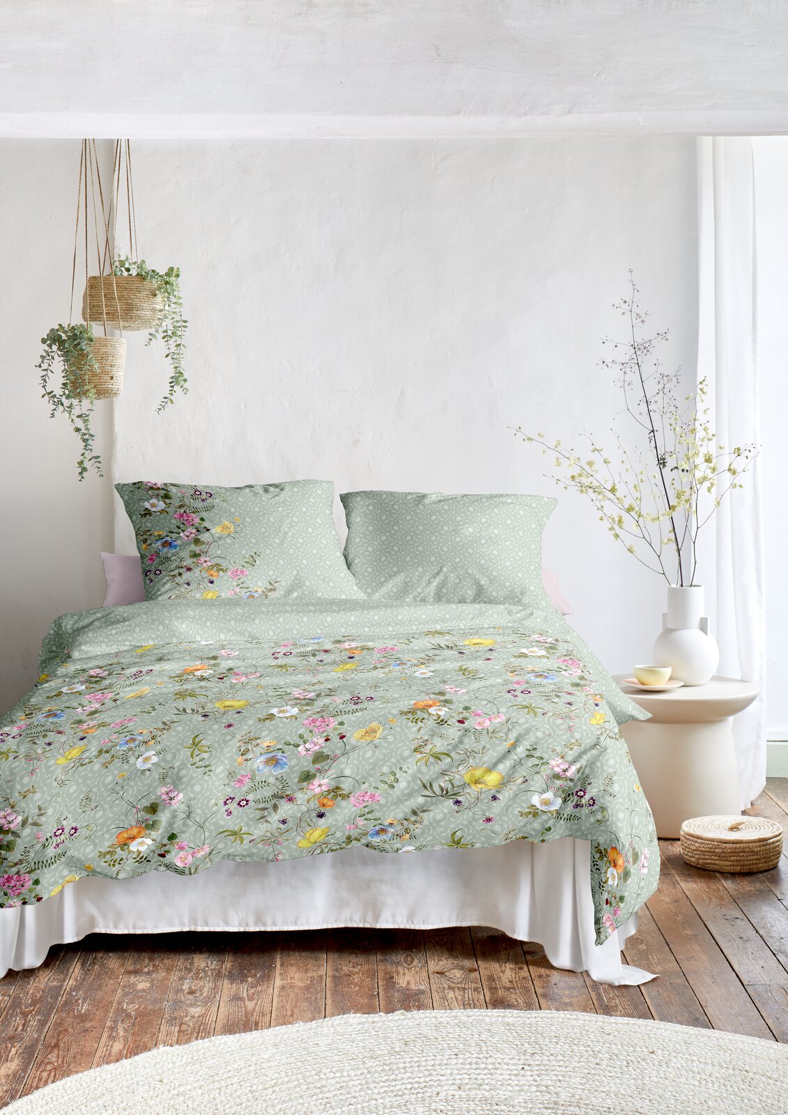 Bettwäsche-Set in sanftem Grün mit buntem Blumenmuster, auf einem Bett in einem hellen Raum mit Holzfußboden und Pflanzen, aus der Frontalperspektive.