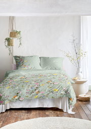 Bettwäsche-Set in sanftem Grün mit buntem Blumenmuster, auf einem Bett in einem hellen Raum mit Holzfußboden und Pflanzen, aus der Frontalperspektive.