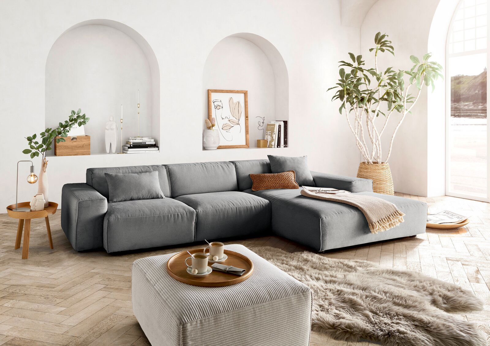 Lounge Handmade Ecksofa LH-EDINA Graue Rundecke in modernem Wohnzimmer mit hellen Wänden, Holzboden und großen Fenstern. Perspektive von vorne links.