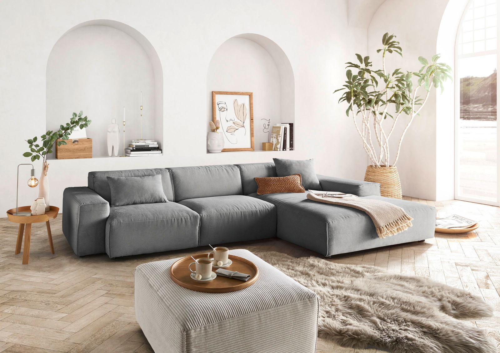Graue Rundecke in modernem Wohnzimmer mit hellen Wänden, Holzboden und großen Fenstern. Perspektive von vorne links.