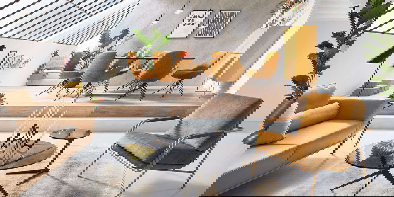 Der Wohnbereich ist hell und stilvoll eingerichtet. Ein bequemer gelber Sessel und ein Sofa laden zum Entspannen ein. Die schwarzen Couchtische haben eine moderne Form. Im erhöhten Essbereich stehen gelbe Stühle um einen runden Tisch. Große Fenster lassen