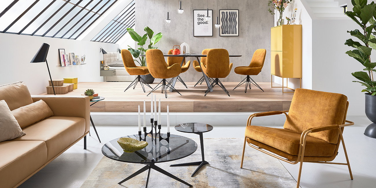 Der Wohnbereich ist hell und stilvoll eingerichtet. Ein bequemer gelber Sessel und ein Sofa laden zum Entspannen ein. Die schwarzen Couchtische haben eine moderne Form. Im erhöhten Essbereich stehen gelbe Stühle um einen runden Tisch. Große Fenster lassen