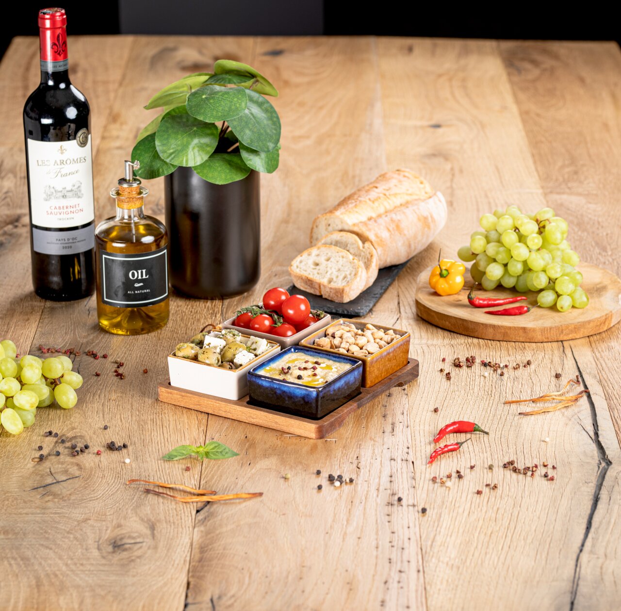 Akazienbrett mit vier Servierschalen, gefüllt mit Oliven, Tomaten, Nüssen und Dip, auf einem Holztisch mit Weinflasche, Ölflasche, Brot und Trauben. Perspektive von oben.