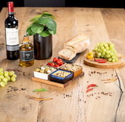 Akazienbrett mit vier Servierschalen, gefüllt mit Oliven, Tomaten, Nüssen und Dip, auf einem Holztisch mit Weinflasche, Ölflasche, Brot und Trauben. Perspektive von oben.