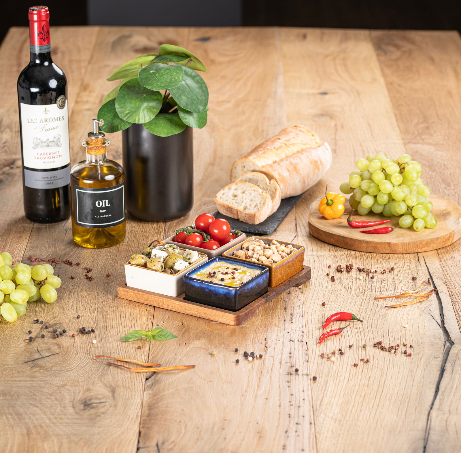 Akazienbrett mit vier Servierschalen, gefüllt mit Oliven, Tomaten, Nüssen und Dip, auf einem Holztisch mit Weinflasche, Ölflasche, Brot und Trauben. Perspektive von oben.
