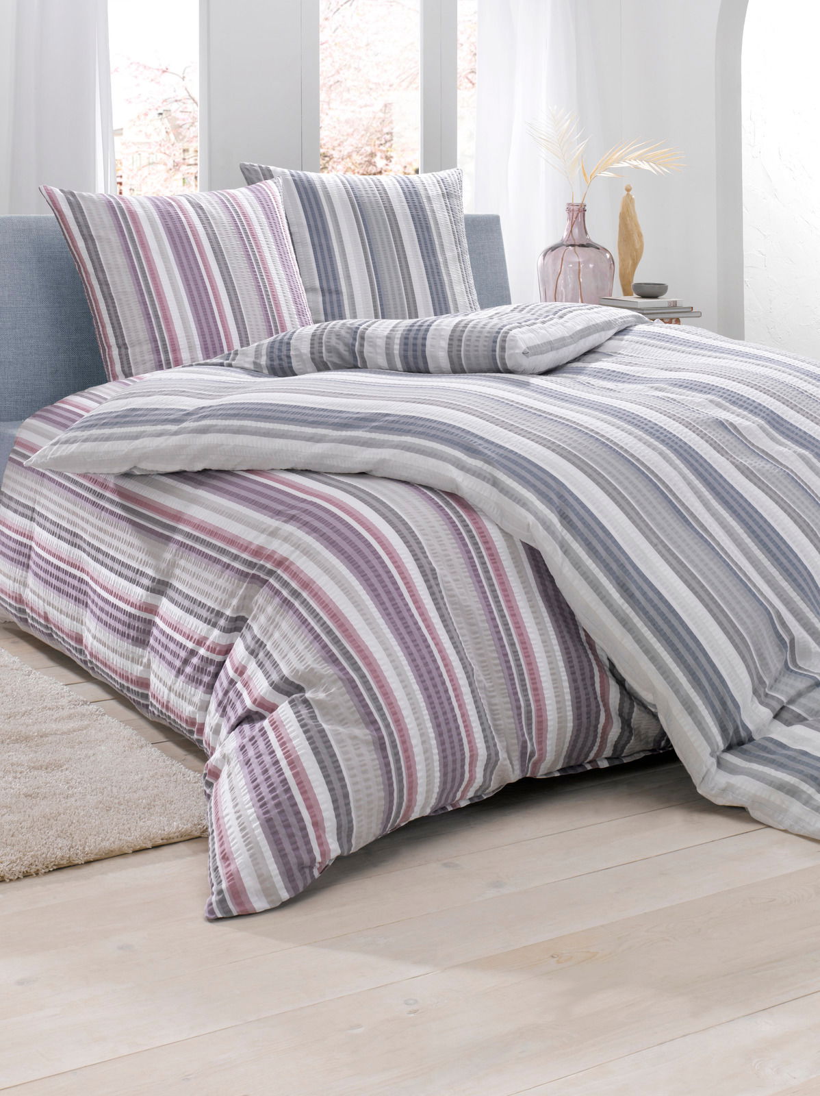 Bettwäsche-Set mit Streifenmuster in Grau- und Lilatönen, bestehend aus einem Bettbezug und zwei Kissenbezügen, auf einem Bett in einem hellen, modernen Schlafzimmer, seitliche Perspektive.