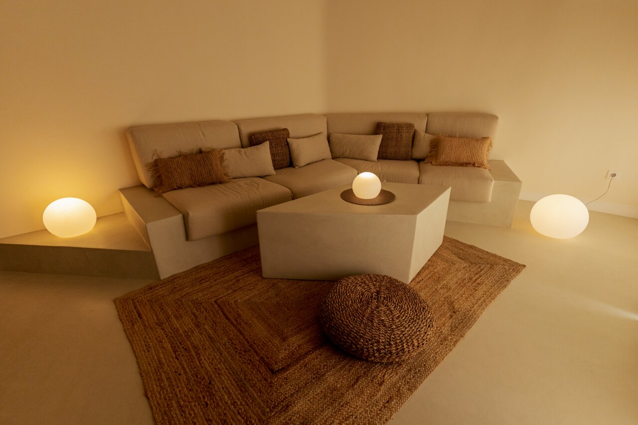 Gezellige, minimalistische zithoek met een beige hoekbank en kussens, een kubusvormige salontafel, een jute tapijt en een ronde staande lamp met warm licht, gefotografeerd vanuit een licht verhoogd vooraanzicht.