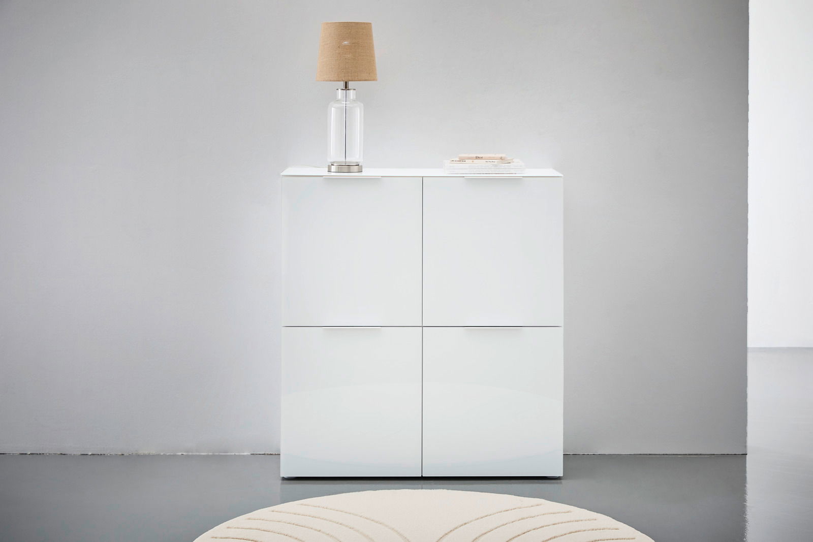 Weißes Highboard mit vier Türen, darauf eine Tischlampe und Bücher, frontale Ansicht.