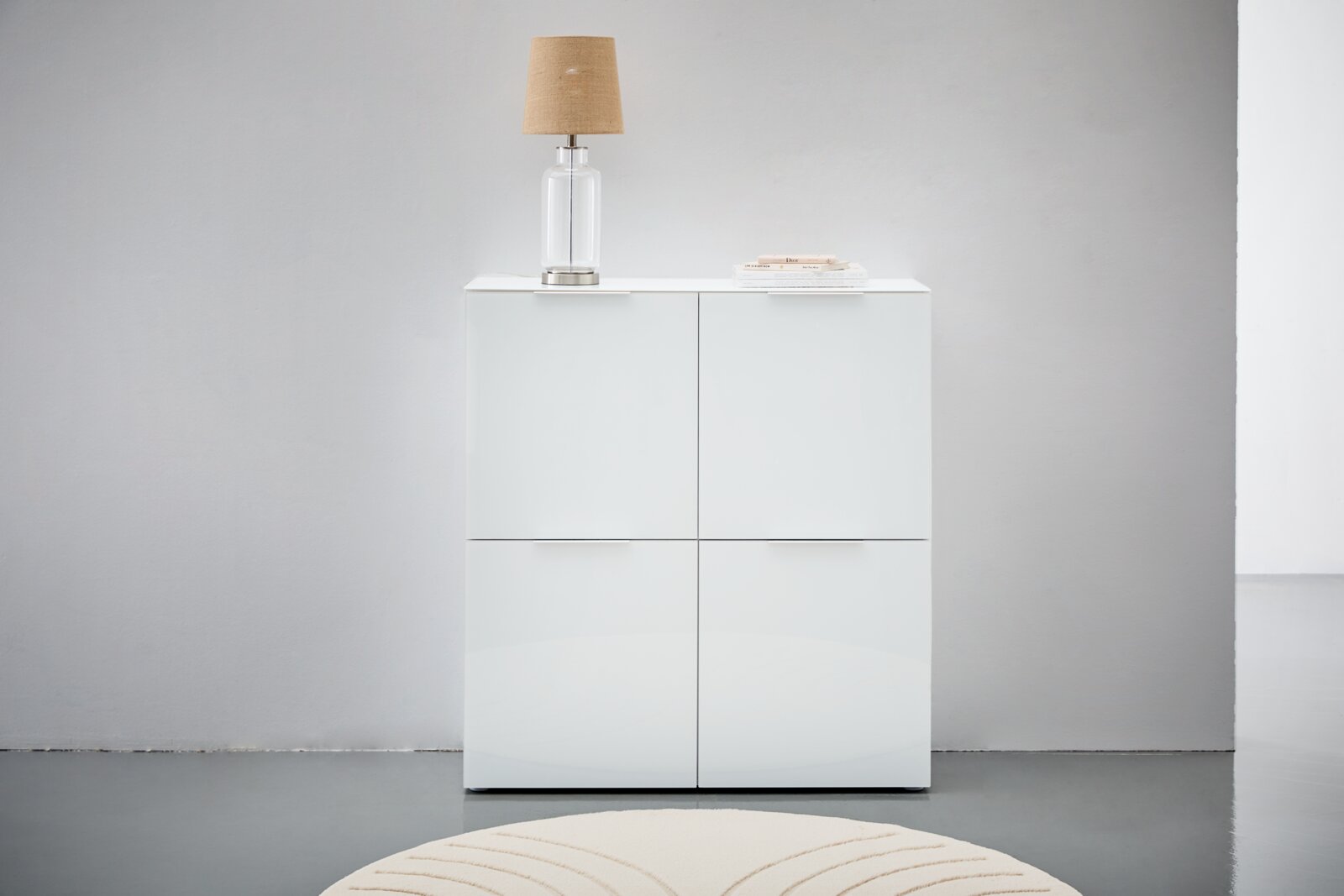 Weißes Highboard mit vier Türen, darauf eine Tischlampe und Bücher, frontale Ansicht.