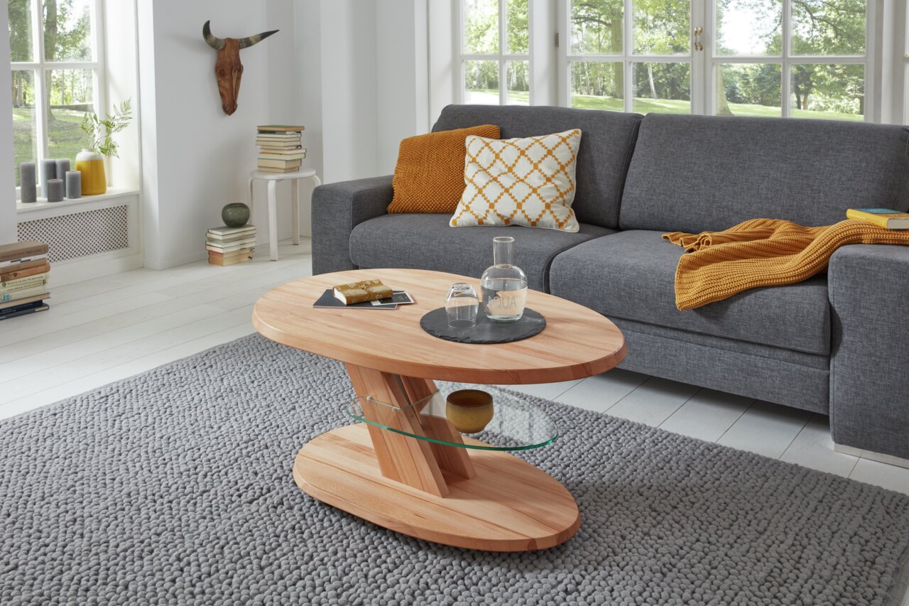 Moderner Couchtisch aus Holz mit Glasablage, in einem gemütlichen Wohnzimmer mit grauem Sofa und großen Fenstern, seitliche Perspektive.