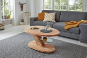 Moderner Couchtisch aus Holz mit Glasablage, in einem gemütlichen Wohnzimmer mit grauem Sofa und großen Fenstern, seitliche Perspektive.