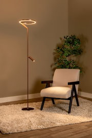 Moderne LED-Stehlampe in Bronze/Kupferoptik mit ringförmigem Leuchtkopf und kleinem Leselicht, warmweiß leuchtend neben einem cremefarbenen Sessel auf Teppich; perspektive: frontale Ansicht leicht von links.