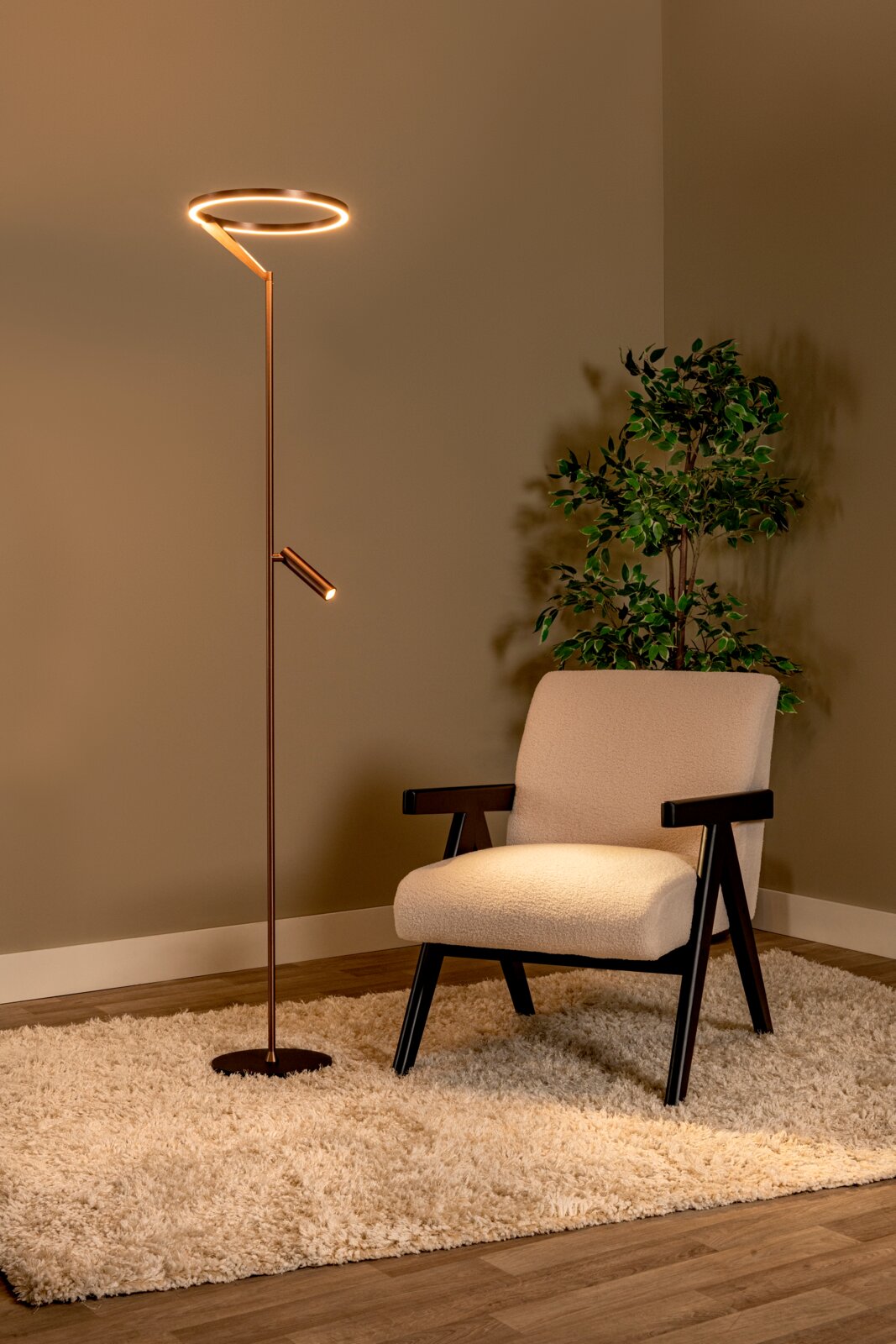 Moderne LED-Stehlampe in Bronze/Kupferoptik mit ringförmigem Leuchtkopf und kleinem Leselicht, warmweiß leuchtend neben einem cremefarbenen Sessel auf Teppich; perspektive: frontale Ansicht leicht von links.