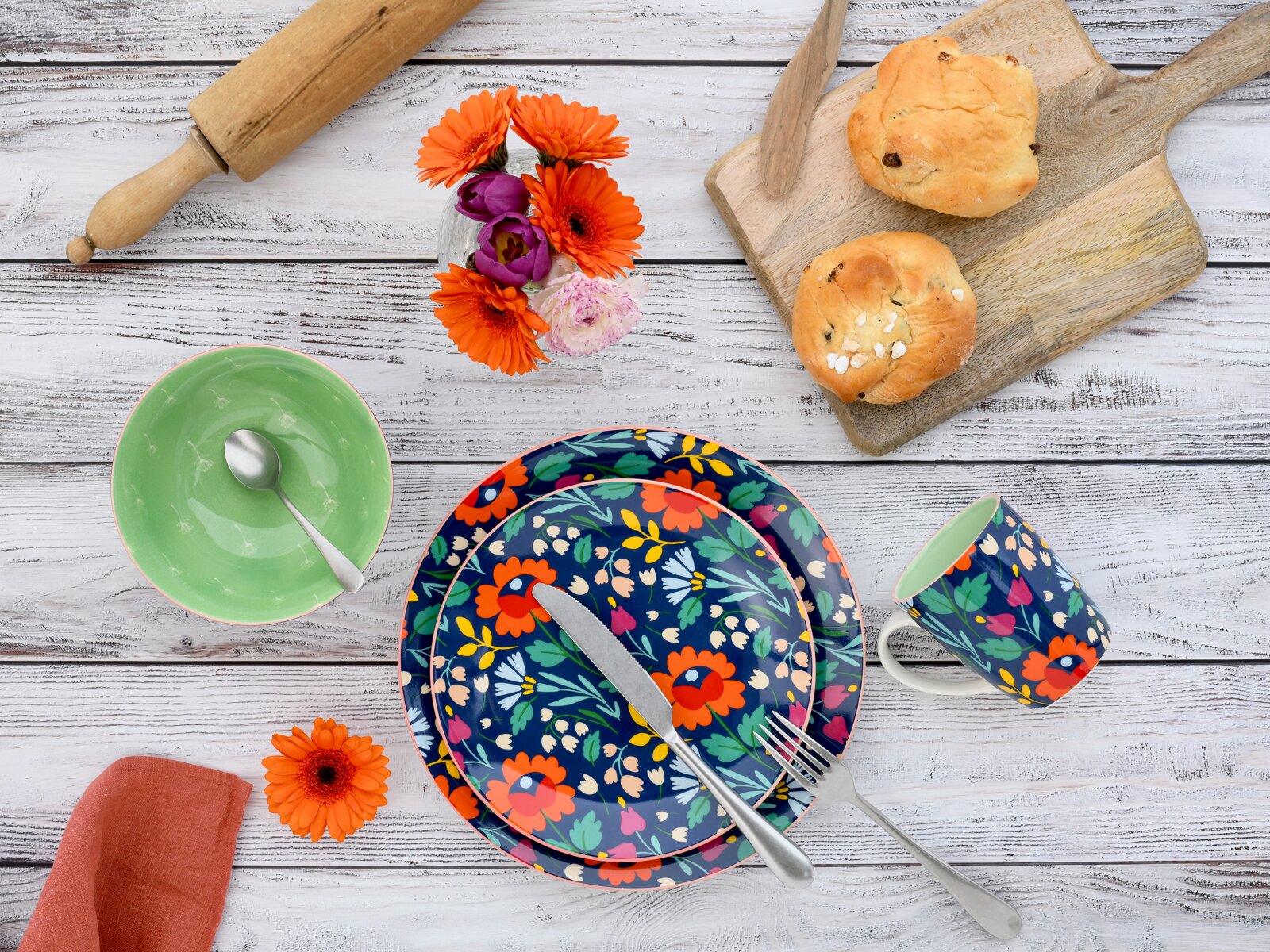 Draufsicht auf farbenfrohes Geschirr-Set mit floral gemustertem Teller und Becher, grüner Schale mit Löffel, Besteck, Holzbrett mit Brötchen, Nudelholz und Blumen auf weißer Holztischplatte