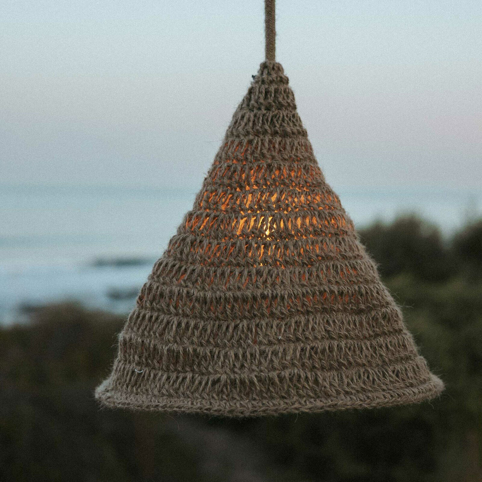 Outdoor-Hängeleuchte aus geflochtenem Material in Kegelform, beleuchtet bei Dämmerung mit Meerblick im Hintergrund, seitliche Perspektive