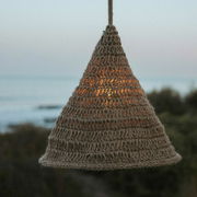 Outdoor-Hängeleuchte aus geflochtenem Material in Kegelform, beleuchtet bei Dämmerung mit Meerblick im Hintergrund, seitliche Perspektive