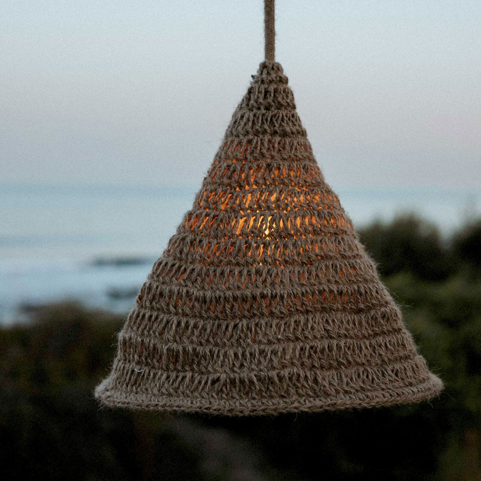 Outdoor-Hängeleuchte aus geflochtenem Material in Kegelform, beleuchtet bei Dämmerung mit Meerblick im Hintergrund, seitliche Perspektive