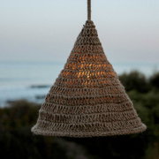 Outdoor-Hängeleuchte aus geflochtenem Material in Kegelform, beleuchtet bei Dämmerung mit Meerblick im Hintergrund, seitliche Perspektive