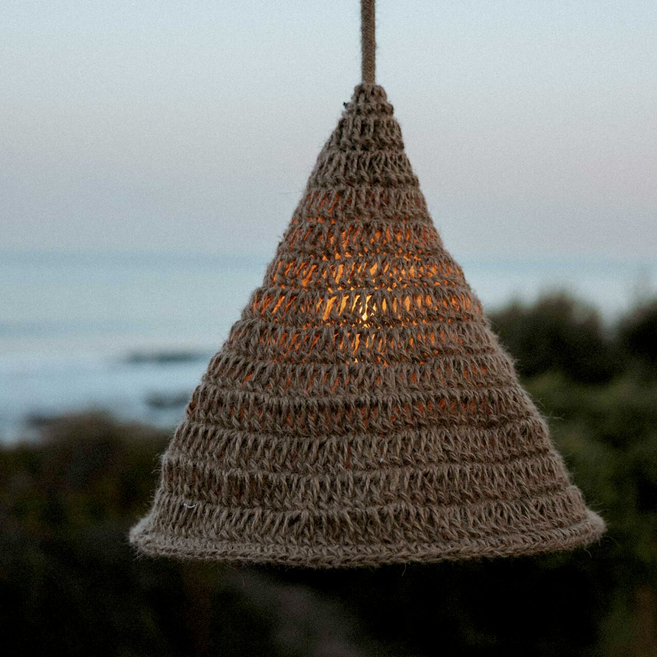 Outdoor-Hängeleuchte aus geflochtenem Material in Kegelform, beleuchtet bei Dämmerung mit Meerblick im Hintergrund, seitliche Perspektive
