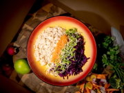 Draufsicht auf eine farbenfrohe Bowl mit Naturreis, glasiertem Lachs, Rotkohl und frischen Sprossen, umgeben von Limette, Radieschen, Petersilie und Gemüse auf einem Holzbrett.