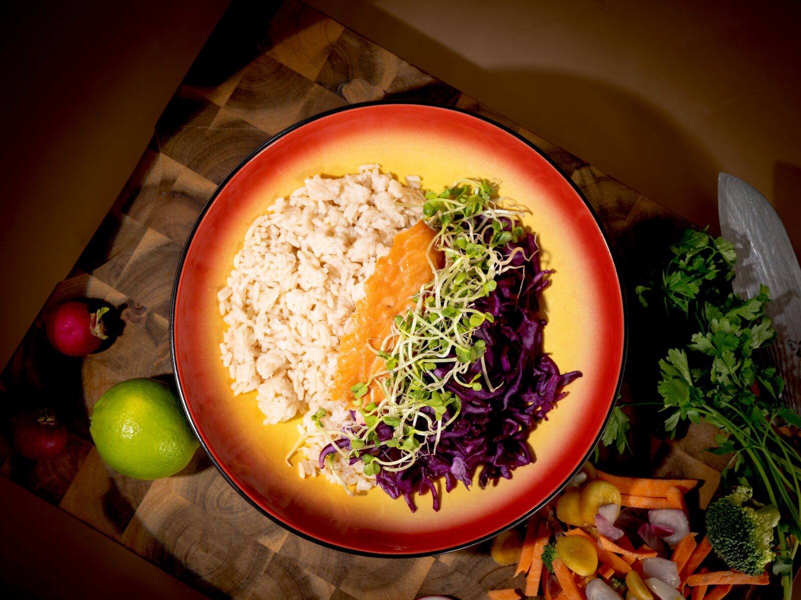 Draufsicht auf eine farbenfrohe Bowl mit Naturreis, glasiertem Lachs, Rotkohl und frischen Sprossen, umgeben von Limette, Radieschen, Petersilie und Gemüse auf einem Holzbrett.