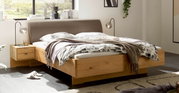 Modern houten bed met een gestoffeerd bruin hoofdeinde en ingebouwde nachtkastjes in een lichte slaapkamer, schuin van voren gefotografeerd vanuit een iets hoger standpunt.