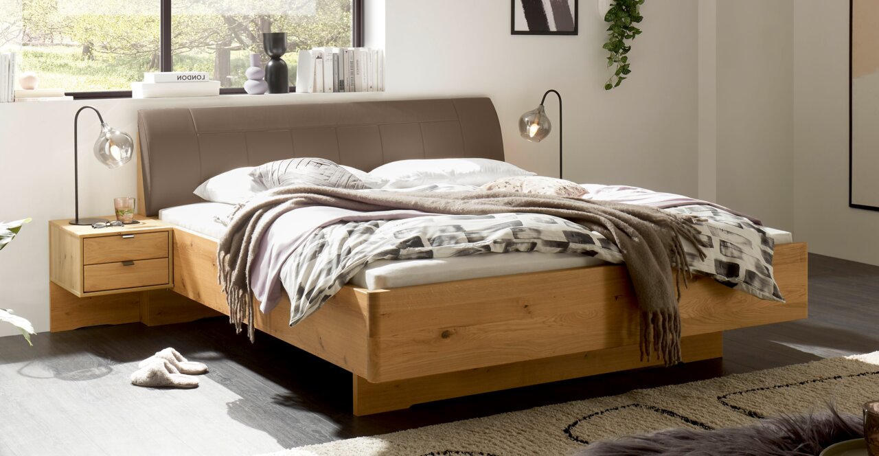 Modern houten bed met een gestoffeerd bruin hoofdeinde en ingebouwde nachtkastjes in een lichte slaapkamer, schuin van voren gefotografeerd vanuit een iets hoger standpunt.
