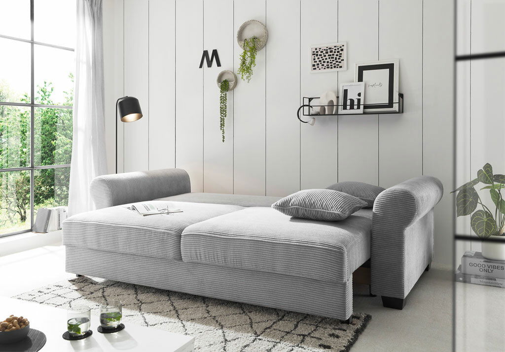 Graues Funktionssofa MAUREEN in einem modernen Wohnzimmer, seitliche Perspektive, mit Kissen und dekorativen Elementen.