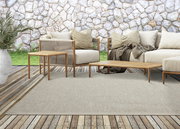 Outdoor-Sitzgruppe mit beigem Teppich, Holztischen und beigen Sofas vor einer Steinmauer, aus der Perspektive von vorne.