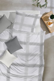 Draufsicht auf ein Bett mit weißer Bettwäsche mit dezentem Streifenmuster, grauen und weißen Kissen sowie einem Holz-Nachttisch mit Pflanze und Gebäck.