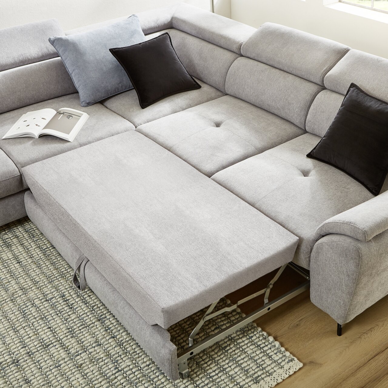 Eckgarnitur in hellem Grau mit ausziehbarem Element, von oben fotografiert, auf einem Teppich stehend, mit dekorativen Kissen.