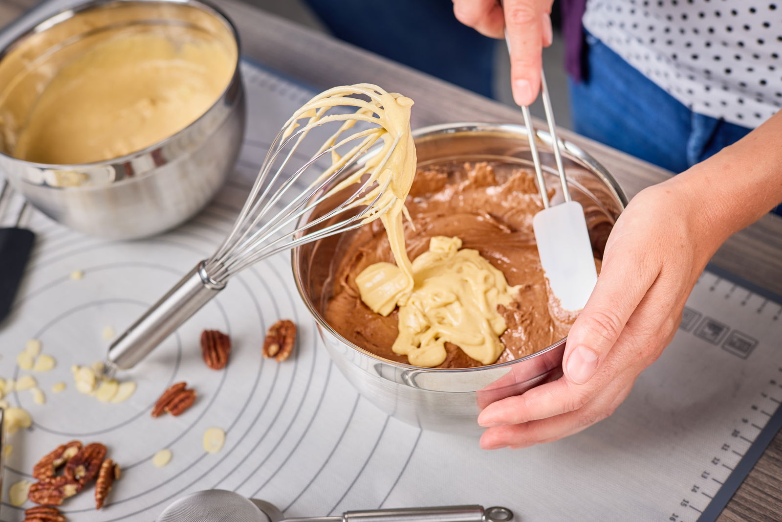Nahaufnahme eines Schneebesens, der in einer Metallschüssel mit Teig verwendet wird. Der Schneebesen ist mit Teig bedeckt, während eine Hand die Schüssel hält und eine andere Hand einen Spatel benutzt. Perspektive von oben.