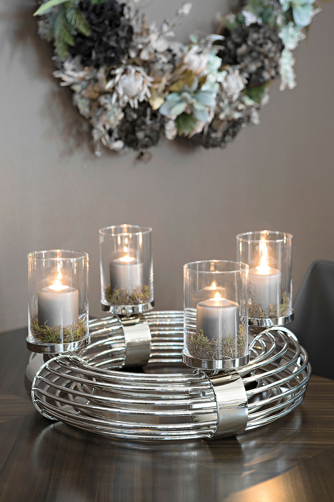 Metallischer Kranz mit vier brennenden Kerzen in Glaszylindern, auf einem Tisch platziert, aus seitlicher Perspektive fotografiert.