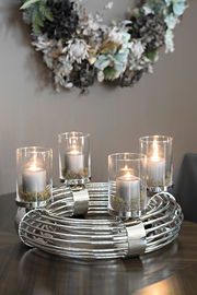 Metallischer Kranz mit vier brennenden Kerzen in Glaszylindern, auf einem Tisch platziert, aus seitlicher Perspektive fotografiert.