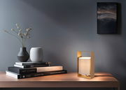 Moderne Tischleuchte mit rechteckigem, goldfarbenem Rahmen und zylindrischem, weißem Lampenschirm auf einem Holztisch, seitliche Perspektive.