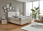 Een modern boxspringbed in een stijlvol ingerichte slaapkamer, gezien vanuit de zijkant, omringd door decoratieve elementen en grote ramen.