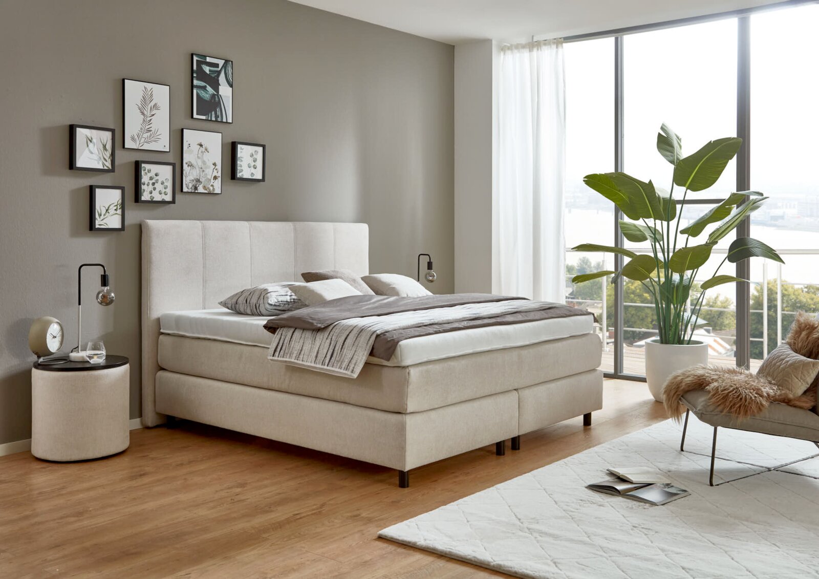 Een modern boxspringbed in een stijlvol ingerichte slaapkamer, gezien vanuit de zijkant, omringd door decoratieve elementen en grote ramen.