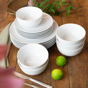 Weißes 18-teiliges Geschirrset auf einem Holztisch, bestehend aus Tellern und Schalen, mit grünen Limetten und Pflanzen im Hintergrund, aus der Vogelperspektive fotografiert.