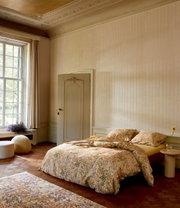Gemütliches Schlafzimmer mit floral gemusterter Bettwäsche in einem eleganten Raum mit großen Fenstern, seitliche Perspektive.