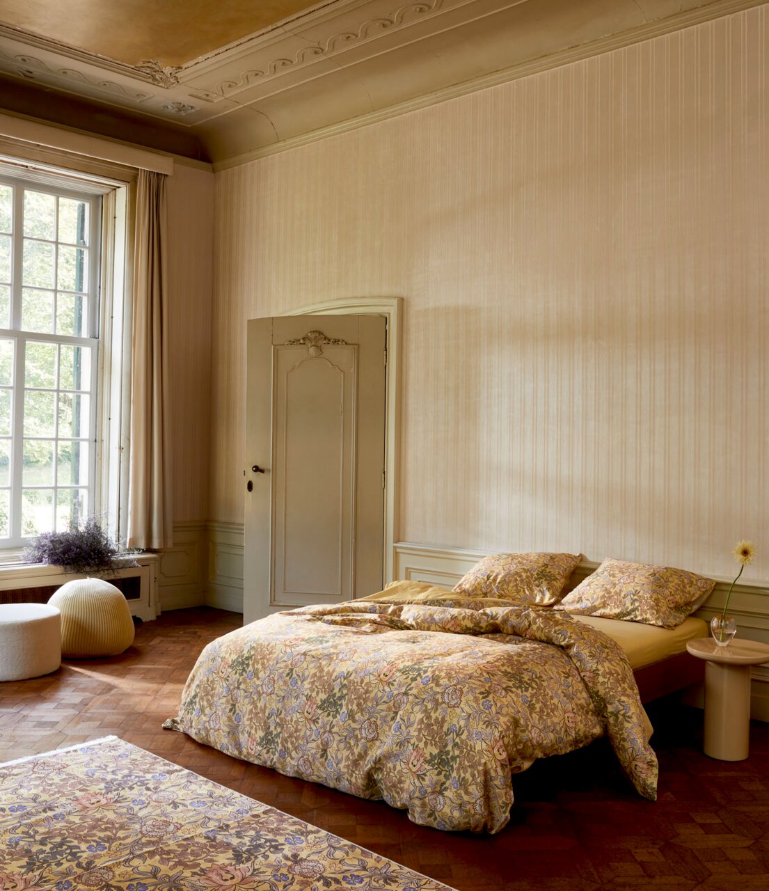 Gemütliches Schlafzimmer mit floral gemusterter Bettwäsche in einem eleganten Raum mit großen Fenstern, seitliche Perspektive.