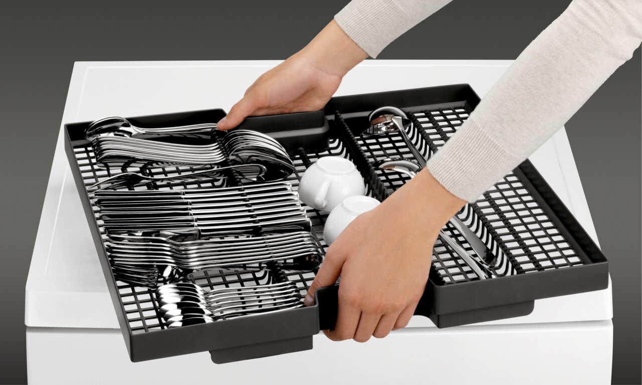Nahaufnahme eines Besteckkorbs in einem Einbaugeschirrspüler, der von zwei Händen herausgezogen wird. Der Korb ist mit Besteck und zwei kleinen Tassen gefüllt.