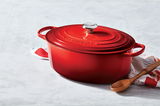 Roter ovaler Bräter mit Deckel und Griffen, seitliche Perspektive, auf einem hellen Untergrund mit Holzlöffel und Geschirrtuch.